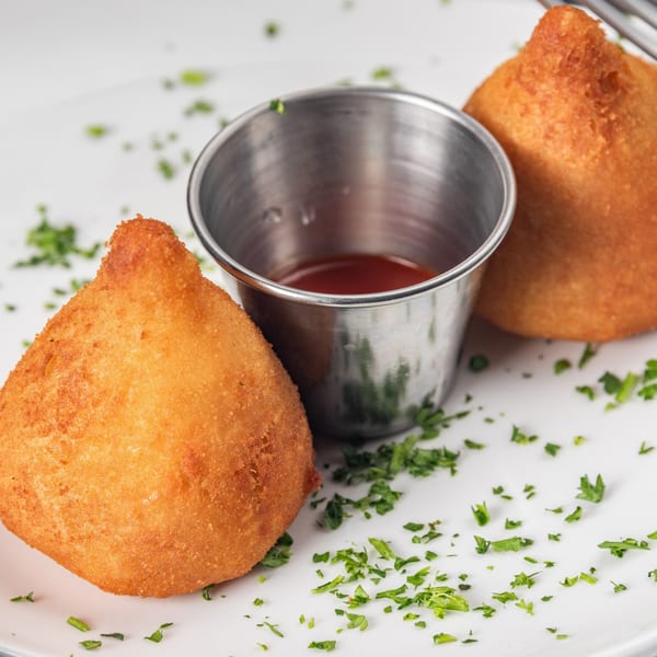 Two golden-brown coxinha pastries with a small metal cup of red sauce and parsley on a white plate.