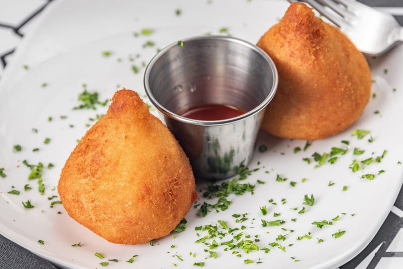 Two golden-brown coxinha pastries with a small metal cup of red sauce and parsley on a white plate.