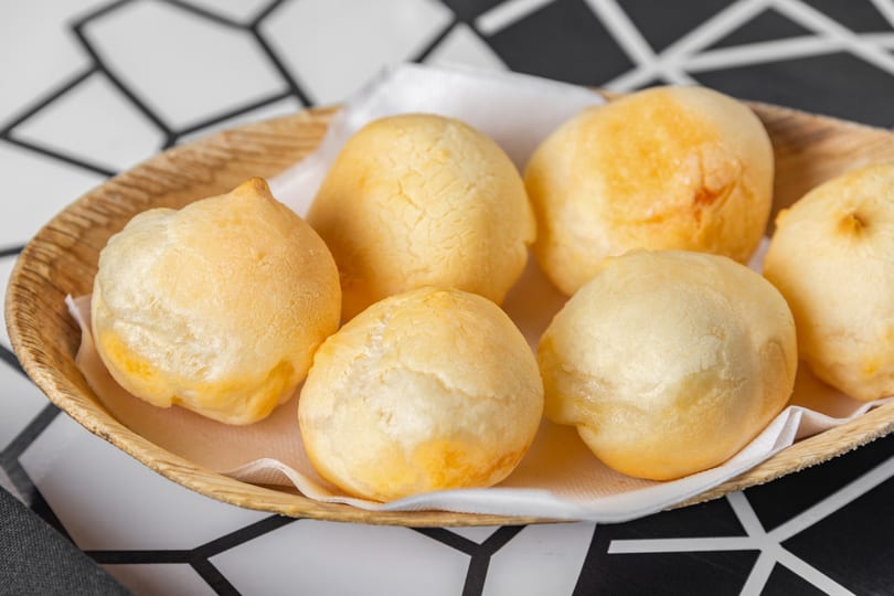Golden-brown Brazilian cheese bread (pão de queijo) served in a woven basket on a patterned tablecloth.