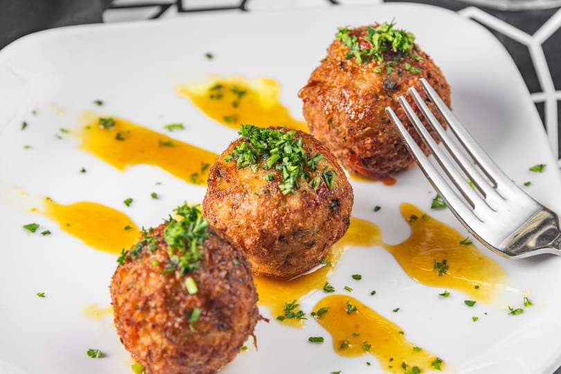 Three golden-brown meatballs with green herbs and orange sauce on a white plate with a fork.
