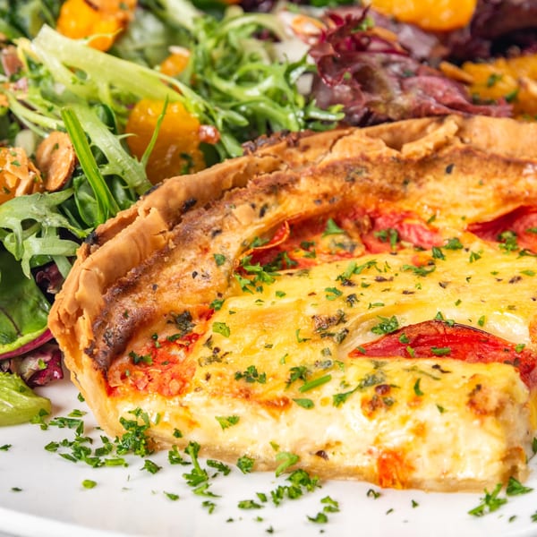 Slice of quiche with tomatoes and herbs, served with a fresh green salad with oranges.